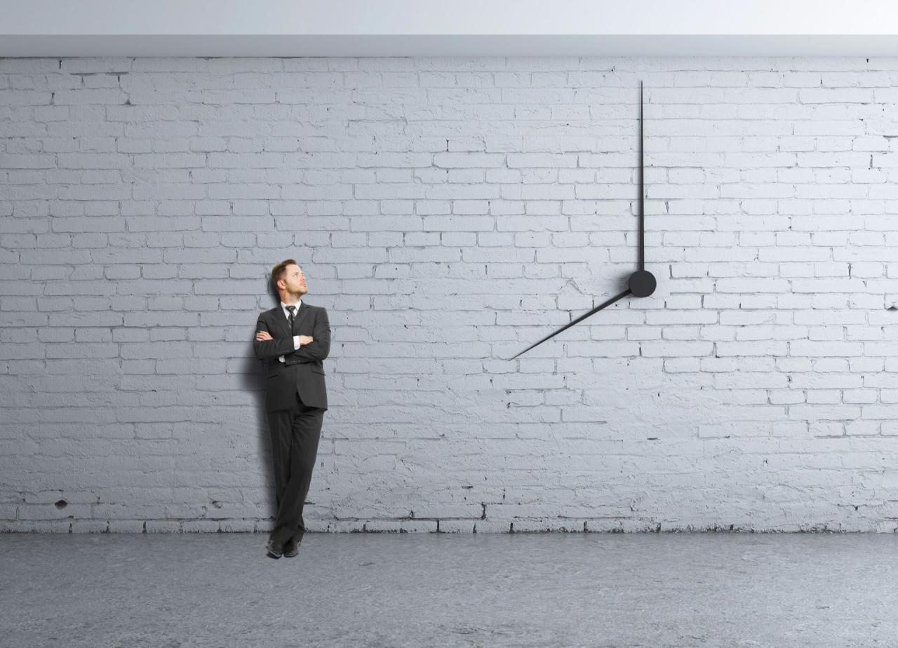 Das Bild zeigt eine weisse Mauer in der zwei große Uhrzeiger integriert sind. Daneben steht einen Mann